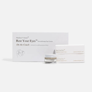 A person in a gray sweater holds an open white box containing MADISON GREEN’s “REST YOUR EYES ON THE COUCH,” lifting the small jar with a white and gold lid from the box—perhaps an essential oil blend designed for calming eye masks or aromatherapy eye covers.