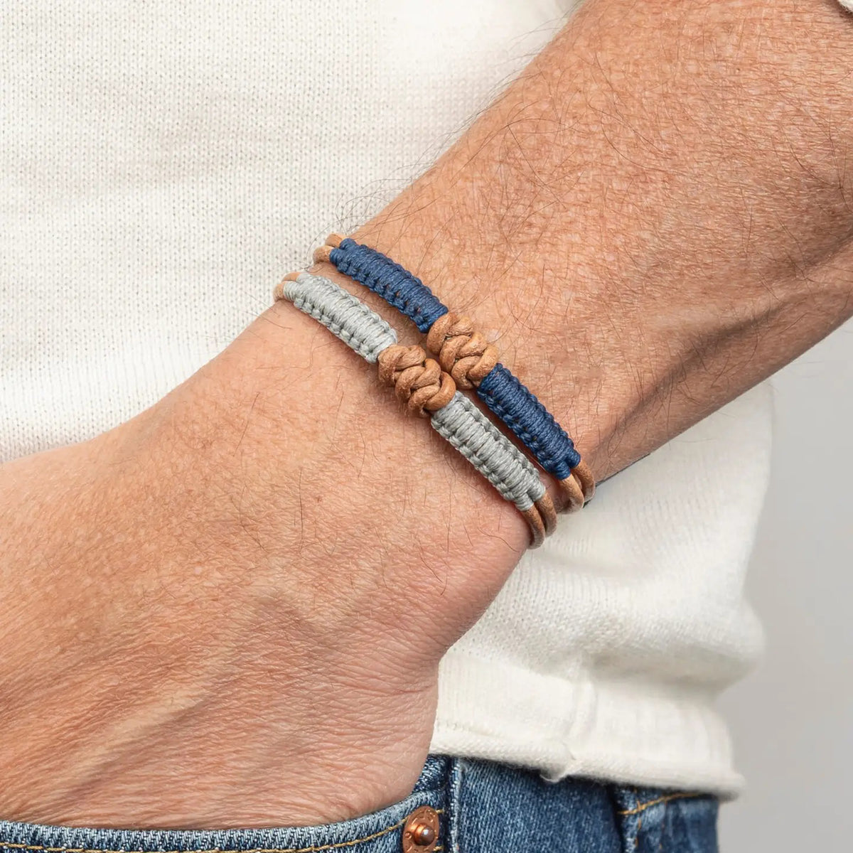 The MAKARLA MEN'S KNOT LEATHER BRACELET ADJUSTABLE IN NAVY is displayed on a white cylindrical stand, labeled by color. This handmade bracelet adds unique style and can be easily adjusted for the perfect fit.
