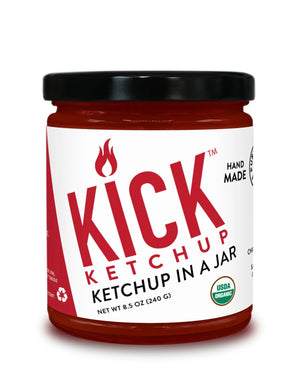 A jar of PURE INDIAN FOODS- KICK KETCHUP by PURE INDIAN FOODS features a black lid, white label with bold red text, flame icon, and "Ketchup in a Jar." This spicy ketchup is handmade with chipotle morita and is USDA Organic.