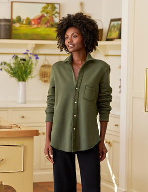 In a sunny, rustic kitchen, a woman smiles while wearing the FRANK & EILEEN EILEEN Relaxed Button Up in Triple Fleece Caper, paired with black wide-leg pants and sandals. She touches her hair as sunlight fills the cozy space.