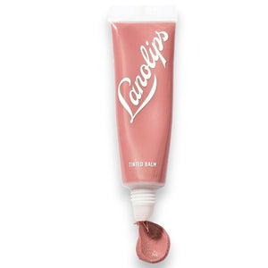 Two arms display swatches of four LANOLIPS TINTED LANOLIN LIP BALM shades—Rose, Rhubarb, Perfect Nude, and Spice—demonstrating the color differences on various skin tones against a pale blue background. The collection features options ideal for anyone wanting a nude lip tint or the nourishing benefits of a LANOLIPS lanolin lip moisturizer.