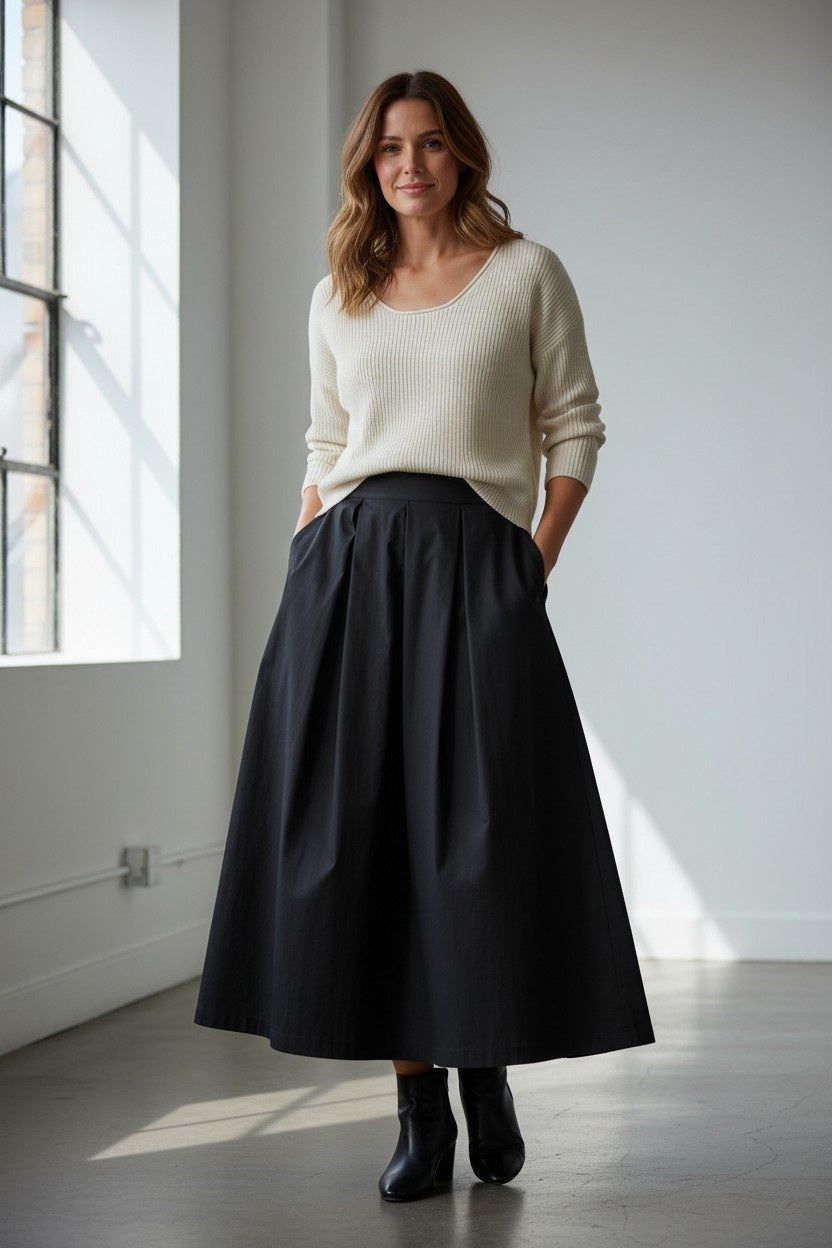 A woman stands indoors wearing a cream sweater, black ankle boots, and the VICARIO CINQUE - GORIZIA MIDI SKIRT IN BLACK TAFFETA. With her hands in her skirt pockets and a soft smile on her face, she is illuminated by sunlight streaming through large windows on the left, highlighting the elegant statement skirt from VICARIO CINQUE.