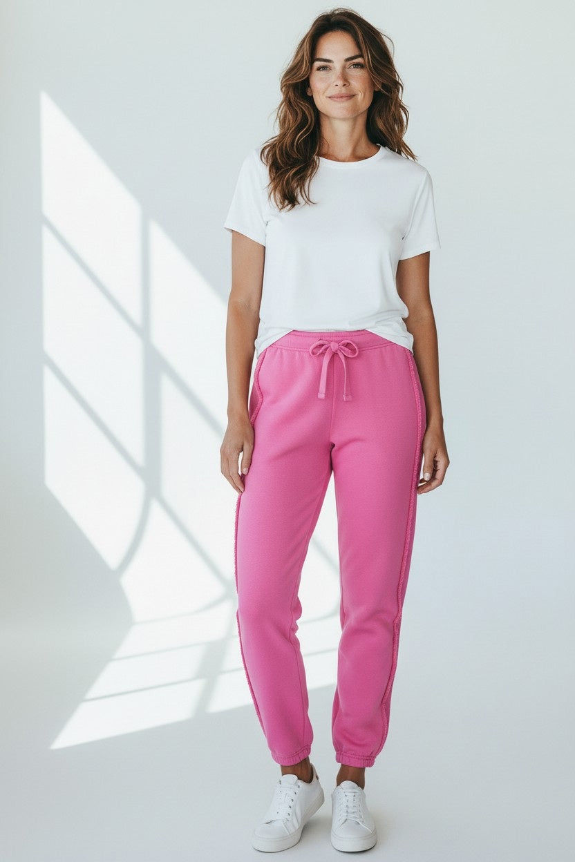 A woman with wavy brown hair smiles in a bright room, wearing FRANK & EILEEN's SOPHIE Italian Sweatpant in Lotus—a 100% cotton, high-rise bright pink jogger—paired with a white tee and sneakers against a plain white background.