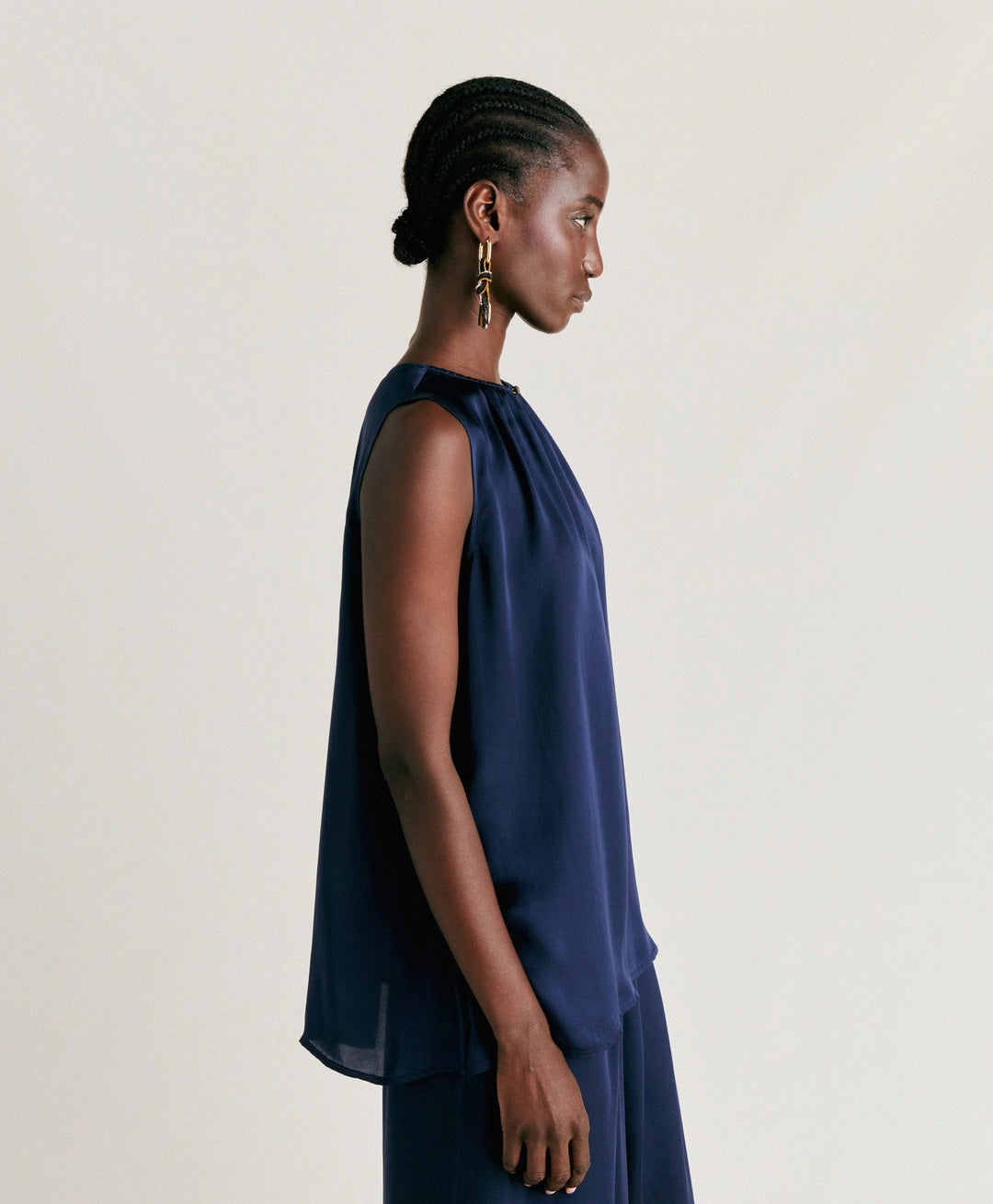 A person stands against a plain background wearing the MOMONI - CRIS SLEEVELESS TOP, a loose-fitting, round neck top in an elegant navy blue. They are also dressed in matching wide-leg pants crafted from pure silk, creating a sophisticated and flowing look. The individual has short, styled hair and is glancing downward with one hand touching their ear.