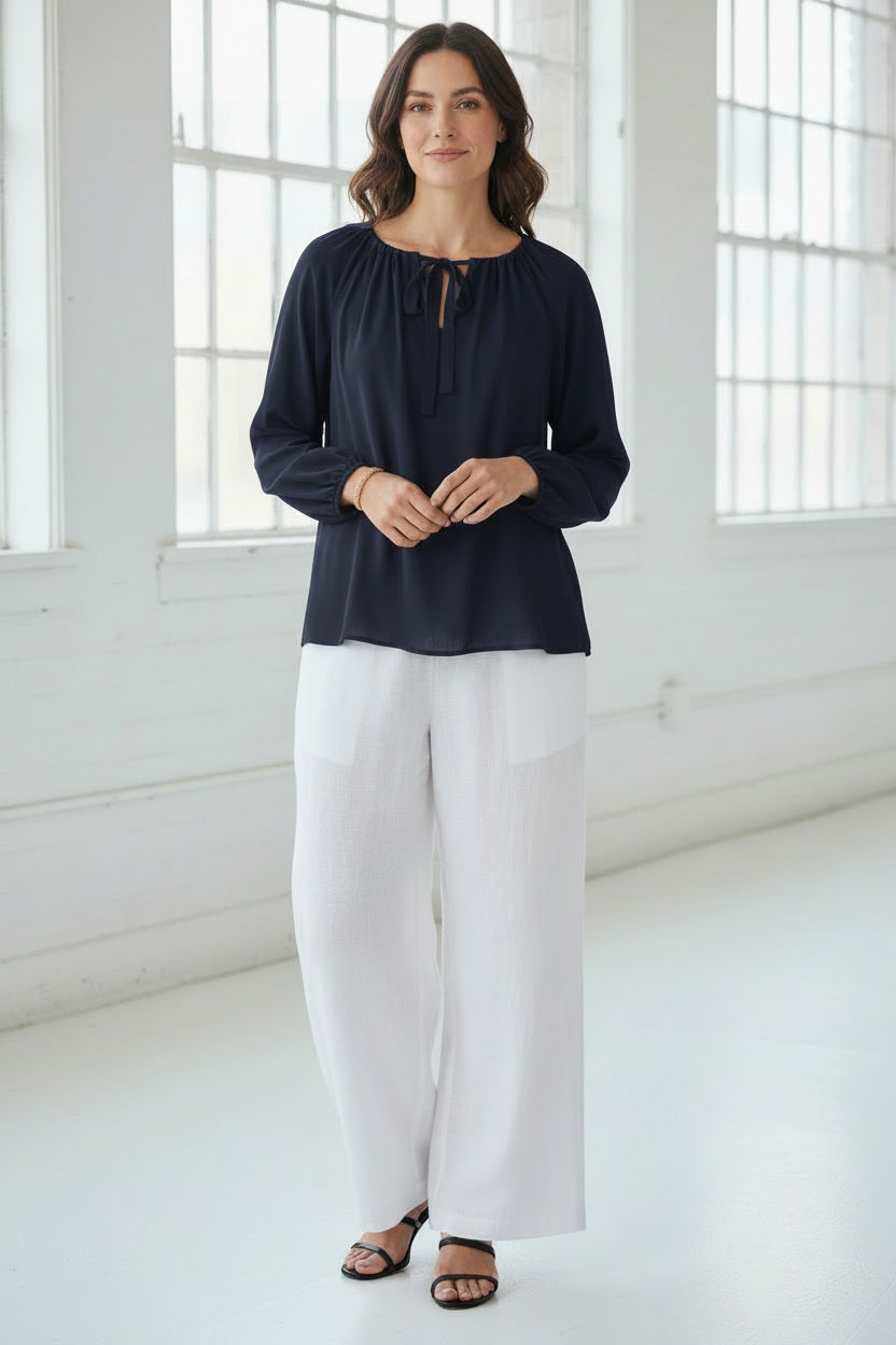A woman in the MEIMEIJ Tie Neck Blouse—loose, long-sleeved, peach-colored washable silk with a neckline tie—paired with white pants, stands against a light textured background. Her face is partially out of frame.