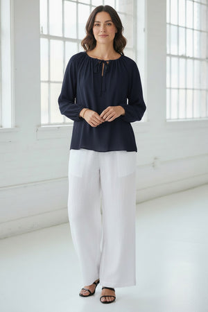 A woman in the MEIMEIJ Tie Neck Blouse—loose, long-sleeved, peach-colored washable silk with a neckline tie—paired with white pants, stands against a light textured background. Her face is partially out of frame.