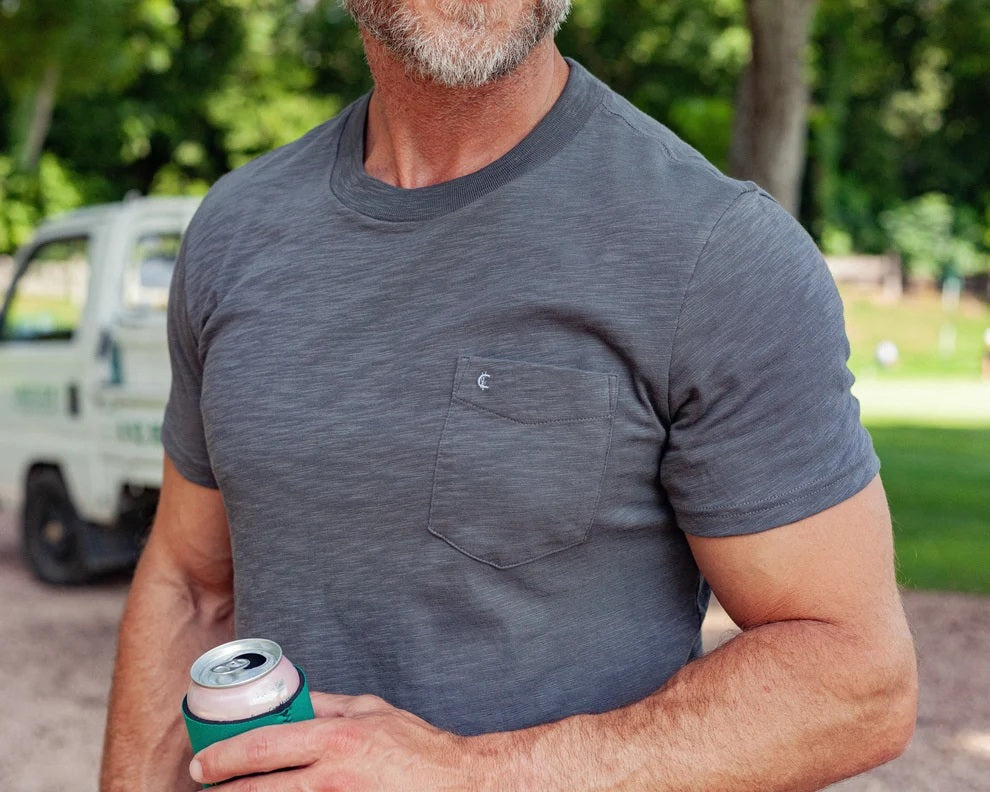 A man with gray hair and a beard, wearing a Criquet Slub Cotton Short Sleeve Pocket T-Shirt in Washed Black and beige pants, holds a green can in his right hand.