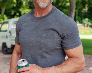 A man with gray hair and a beard, wearing a Criquet Slub Cotton Short Sleeve Pocket T-Shirt in Washed Black and beige pants, holds a green can in his right hand.