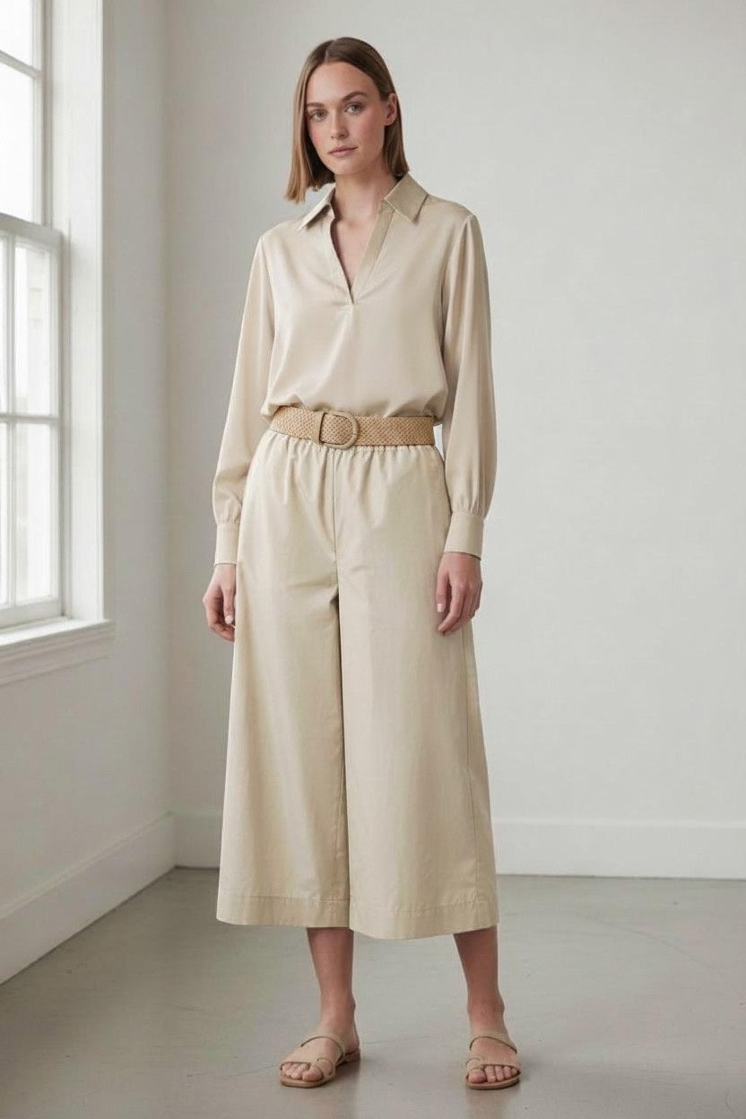 A woman stands indoors in natural light, wearing a beige long-sleeve blouse, sandals, and the VICARIO CINQUE - PAPILO Wide Leg Taffeta Pant in Beige by VICARIO CINQUE. She has straight hair and a neutral expression.