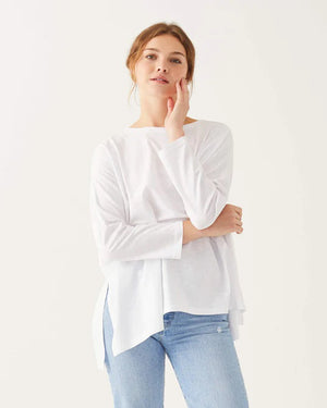 A woman with long, straight hair stands against a white wall, wearing the MERSEA Catalina Slub Tee in blue paired with white pants. The midweight textured cotton tee is from MERSEA. She looks at the camera with a neutral expression.