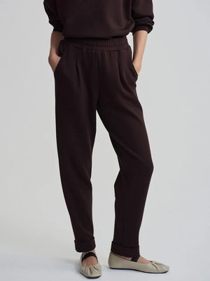 A person seen from behind wearing VARLEY - ROLLED CUFF PANT 25” IN COFFEE BEAN dark brown trousers with rolled cuffs and cream-colored ballet flats.