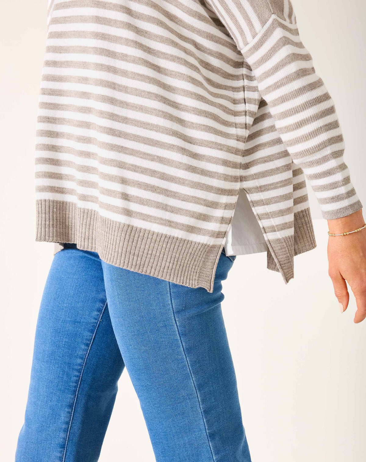A woman with blonde hair wears the MERSEA - CATALINA MODERN CREWNECK SWEATER in gray and white stripes over a white collared shirt and blue jeans, standing against a plain white wall with a slight smile, one arm bent across her body.