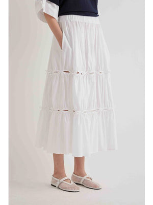A person stands sideways in a MEIMEIJ cotton poplin midi skirt with bow detail and gathered tiers, paired with a black rolled-sleeve top and white mesh ankle-strap shoes. The background is plain and light-colored.