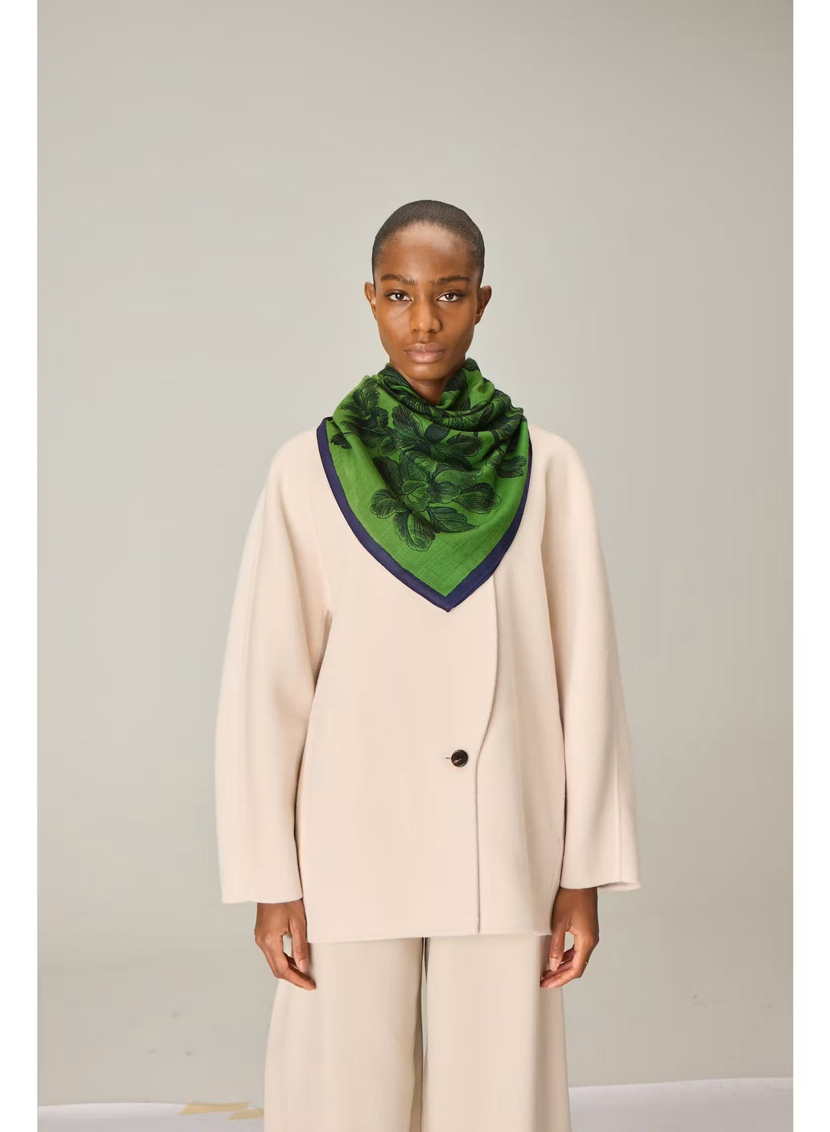 A person wearing a cream-colored suit and the INOUI EDITIONS - ARNORIS WOOL SCARF IN GREEN by INOUI, elegantly draped like a 100% wool shawl made in India, stands facing forward against a plain light background.