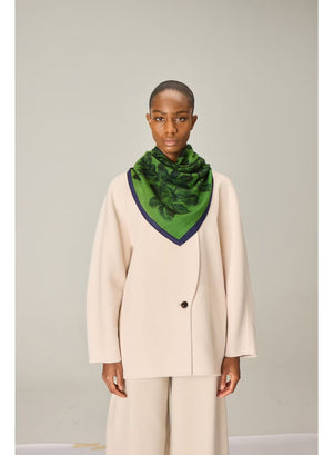 A person wearing a cream-colored suit and the INOUI EDITIONS - ARNORIS WOOL SCARF IN GREEN by INOUI, elegantly draped like a 100% wool shawl made in India, stands facing forward against a plain light background.