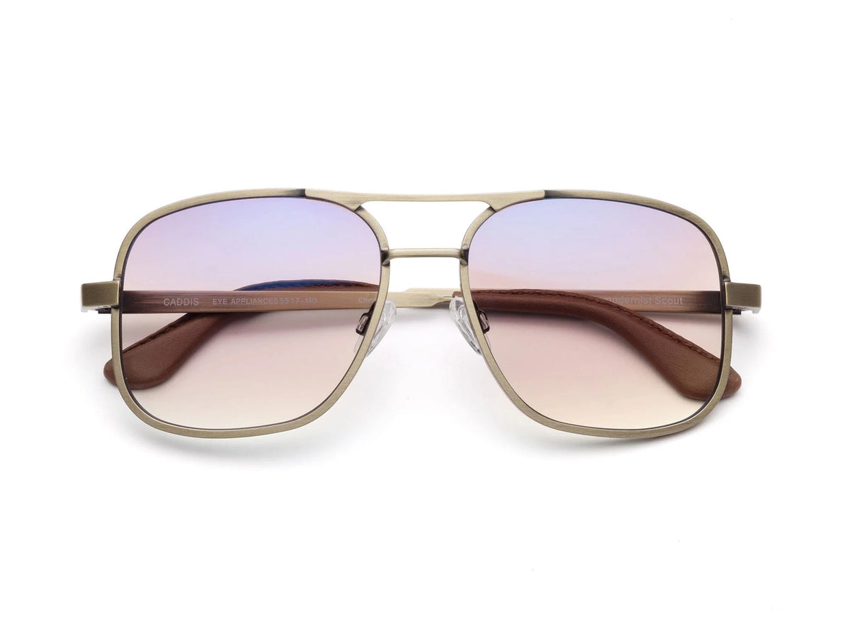 The CADDIS - METAMODERNIST SCOUT READERS by CADDIS are aviator-style sunglasses featuring a slim metal alloy frame and gradient, scratch-resistant lenses that fade from light blue to pink, displayed on a white background.