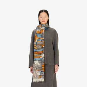 A woman stands facing forward, wearing a grey long-sleeve coat and skirt, black shoes, and the INOUI EDITIONS Stack of Books Wool Scarf in navy, a colorful, hand-printed accessory from INOUI, made in India, on a white background.