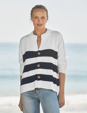 A woman stands on a beach wearing the FRANK & EILEEN Montecito Crew Neck Cardigan in Pure Italian Cotton, featuring navy and white stripes in a relaxed fit with large buttons, paired with light blue jeans. The ocean and sky are softly blurred in the background.