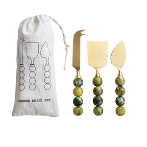 The CREATIVE COOP GOLDTONE CHEESE KNIVES WITH GREEN RESIN BALL HANDLES, SET OF 3, shine atop a cookbook with green pages on a wooden surface—just what you need for your next charcuterie board.