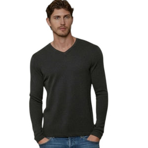 A man stands against a beige wall with his hands in his pockets, wearing the AUTUMN CASHMERE - Cashmere Basic V Neck with Open Bottom in Pepper by AUTUMN CASHMERE, paired with blue jeans and white sneakers.