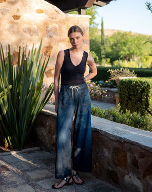 Wearing the MERSEA Londonberry Drawstring Jeans in Night Sky—a pair of wide-leg dark denim jeans with a comfortable drawstring waist—from MERSEA, styled with a cream sleeveless top, a striped shirt draped over the shoulders, and brown sandals, standing against a plain light background.