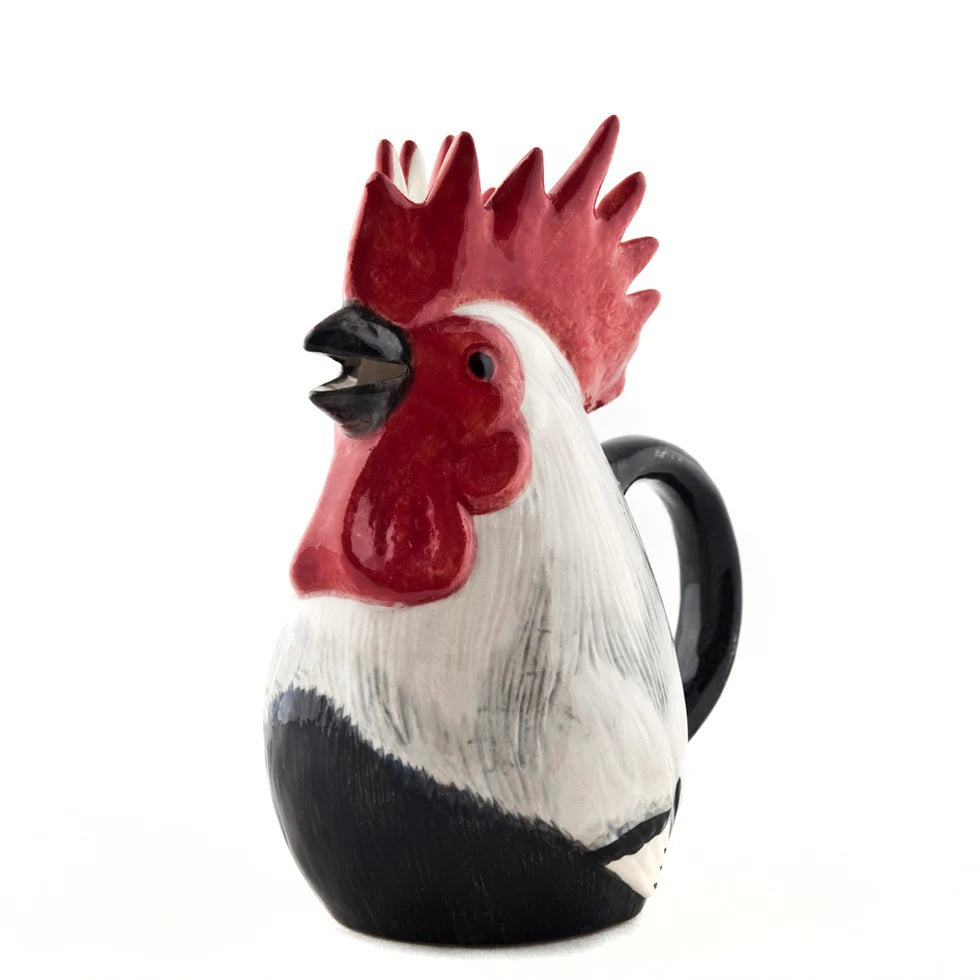 The QUAIL CERAMICS - DORKING ROOSTER JUG by QUAIL DESIGNS, a hand-painted stoneware jug with a red comb and black handle, sits on the table beside a green glass and patterned plates—a charming British tea accessory.