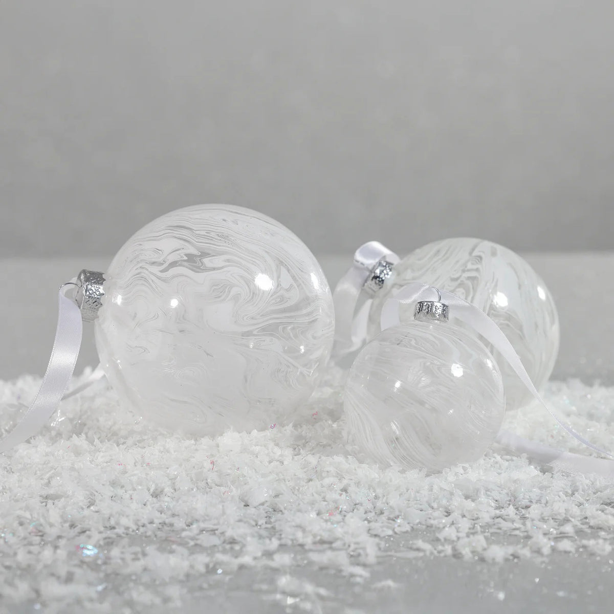 The ZODAX WATERCOLOR GLASS ORNAMENT IN WHITE is a round, marbled 4-inch Christmas ornament suspended by a thin silver ribbon against a plain white background.