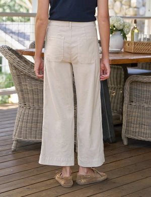 A person wearing the FRANK & EILEEN Limerick Cropped Wide Leg Carpenter Pant in Italian Performance Linen Cement, styled with tan lace-up shoes, stands indoors on a wooden floor with wicker furniture in the background.