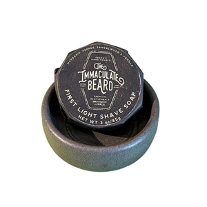 A round tin of THE IMMACULATE BEARD - SHAVE SOAP PUCK IN FIRST LIGHT rests on marble, its label noting mandarin, pepper, sandalwood, and vanilla scents for sensitive skin. Hand-poured by THE IMMACULATE BEARD; net weight 3 oz.