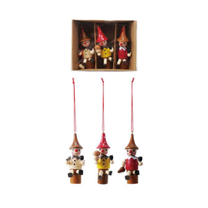 Three CREATIVE COOP WOOD WOODSMEN ORNAMENTS with red hats hang above a boxed set of four similar multi-colored figures, each in its own compartment, all displayed on a speckled white surface.