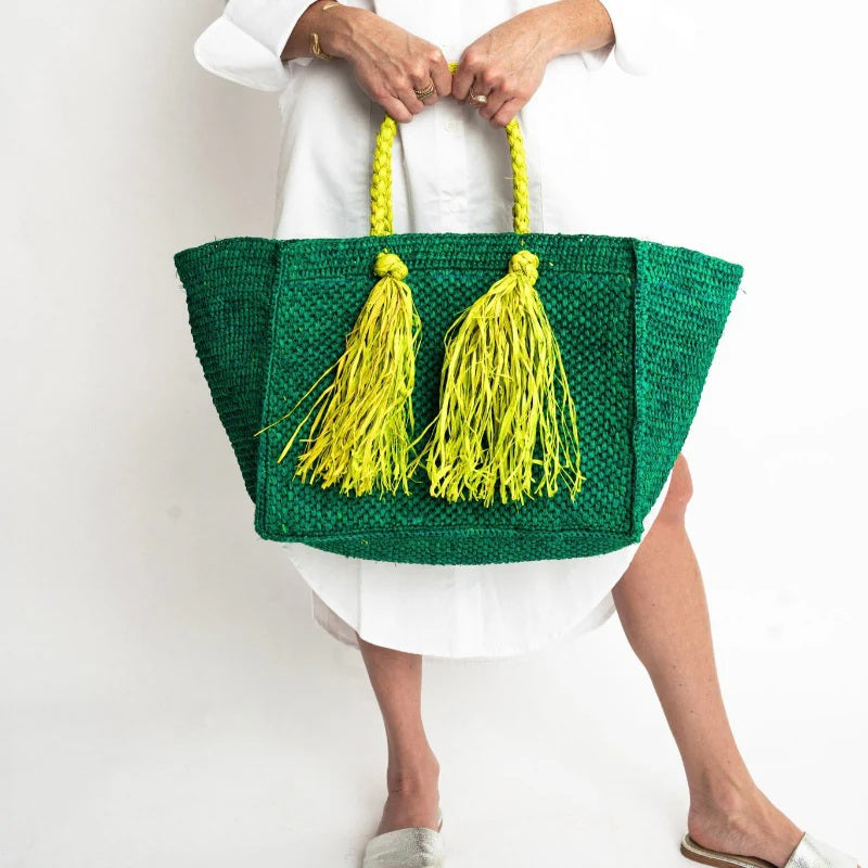 A person in a white dress holds the BALDIZ - CAMILLA RAFFIA BAG IN EMERALD AND LIME, featuring two bright yellow braided handles and oversized yellow tassels, against a plain white background.