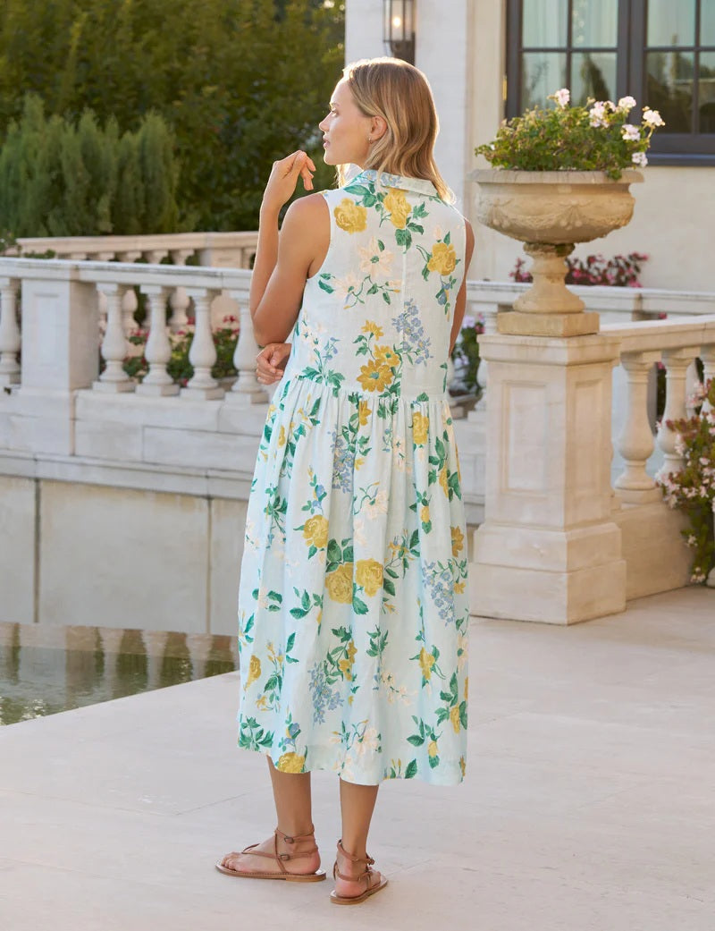 A woman smiles outside near a stone railing and planter, wearing the FRANK &amp; EILEEN Daphne Popover Dress in Italian Linen Summer Scatter. She pairs it with brown sandals; greenery and a reflective water feature are in the background.