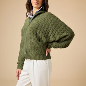 A woman wearing a cream-colored, plush SOFTIES - CABLE KNIT MARSHMALLOW FUNNEL NECK DOUBLE ZIP JACKET with a funnel neck and double zipper, paired with blue jeans.