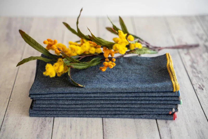 A stack of DOT AND ARMY - CHAMBRAY DINNER NAPKINS rests on a light wooden surface, topped with a sprig of yellow flowers and green leaves.