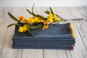 A stack of DOT AND ARMY - CHAMBRAY DINNER NAPKINS rests on a light wooden surface, topped with a sprig of yellow flowers and green leaves.