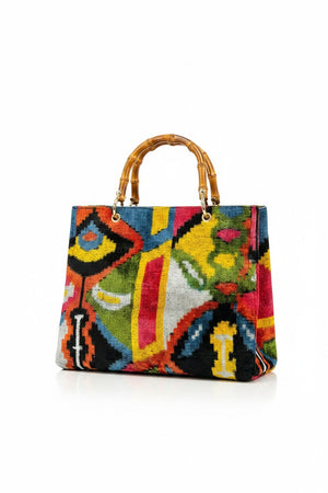 A person in white pants holds the BALDIZ - AVERY BAG IN PALOMA WITH BAMBOO HANDLES, featuring a colorful abstract pattern, against a plain background.