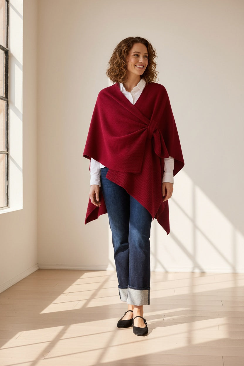 A woman in the MERSEA - Modern Travel Wrap in Wine, featuring a securing loop, pairs it with a white shirt and rolled-up jeans as she walks carrying a large black bag. She wears black flats and her light brown hair is tied back. The background is plain beige.