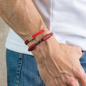 The MAKARLA MEN'S LEATHER ROPE BRACELET IN RED features a handmade design with an adjustable sliding knot closure, stylishly displayed on a white cylindrical stand and labeled with its color.