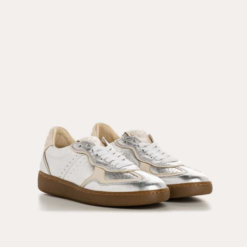 The REQINS - ATHENA METALLIC SNEAKER IN SILVER features a metallic silver finish, white laces, beige heel accents, suede leather details, and a brown rubber sole, displayed in side profile against a plain light background.