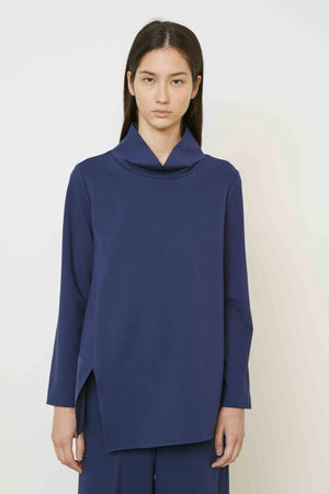 A woman with long brown hair, seen from behind, wears the MEIMEIJ Mixed Media Cowl Neck Top in dark blue with a shiny panel at the bottom and matching pants. She stands against a plain, light background.