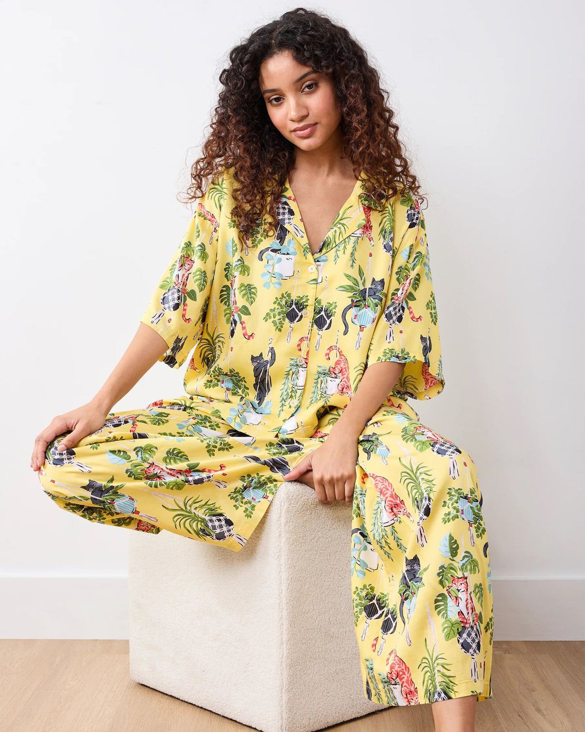 A woman stands barefoot indoors against a plain white wall and light wood floor, wearing the PRINTFRESH - Macrame Mischief Satin Wildest Dream Pajama Set in Sunshine by PRINTFRESH, featuring a loose-fitting eco satin yellow fabric with a colorful floral and cat print.