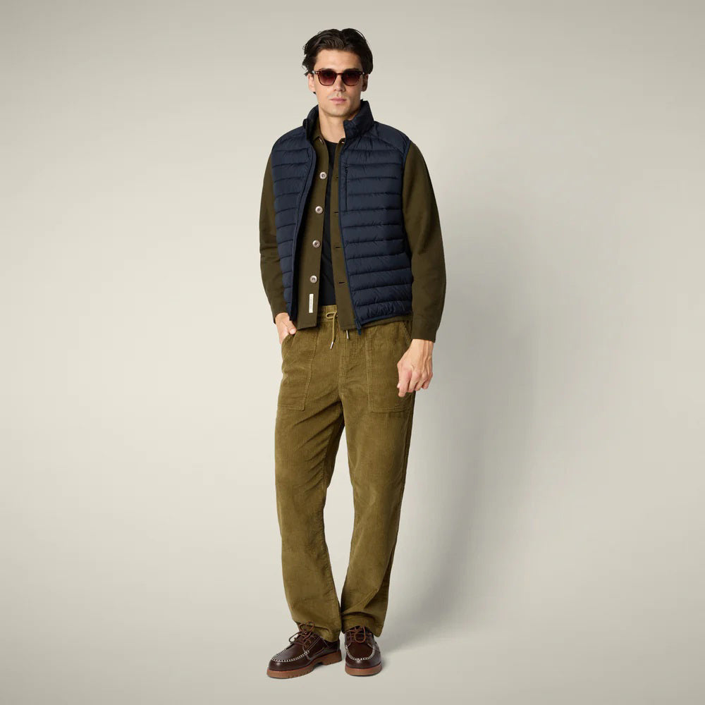 A man wearing the SAVE THE DUCK - RHUS VEST with Recycled Plumtech padding over an olive cardigan, paired with loose-fitting olive pants and brown shoes, stands against a plain light background with his hand in his pocket.