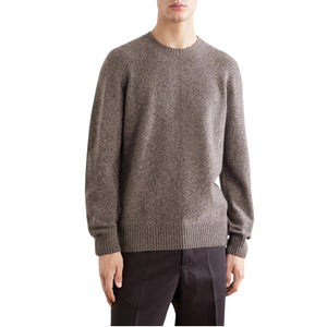 A person wearing the HARTFORD Seamless Wool Crew Pullover in Mushroom, an Italian-made sweater, paired with dark pants, is seated with one arm resting on their knee against a plain light background.