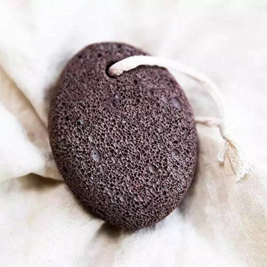 The SMILE BOUTIQUES NATURAL PUMICE STONE FOOT SCRUBBER features a brown, rough, and porous surface ideal for foot care and effective callus removal. It comes with a white rope loop for convenient hanging, as shown against a white background.