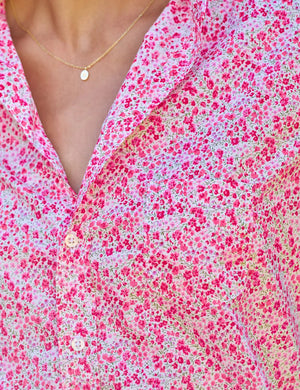 A woman stands on a brick path in a lush garden, wearing the FRANK & EILEEN EILEEN Relaxed Button Up in Liberty Print Pink Flowers, with blue jeans and woven flats. She carries a tote of pink flowers, surrounded by greenery and blooms.