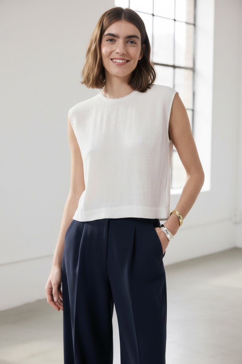 A woman with shoulder-length brown hair smiles, wearing the HARTFORD HOWIE Sleeveless Top by HARTFORD and high-waisted navy trousers, standing in a bright room with large windows.