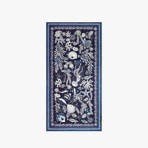 A woman faces forward, wearing a sleeveless cream and navy top with white botanical and animal motifs, accessorized with the INOUI EDITIONS - NEPTUNE SCARF IN NAVY by INOUI, draped loosely around her neck.