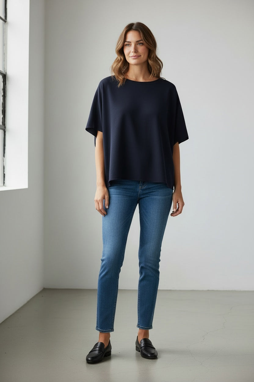 A woman stands indoors wearing the FRANK & EILEEN Olive Original Capelet in British Royal Navy, paired with blue jeans and black loafers. She has wavy hair and is smiling gently, one hand by her side. The room features white walls and a large window.
