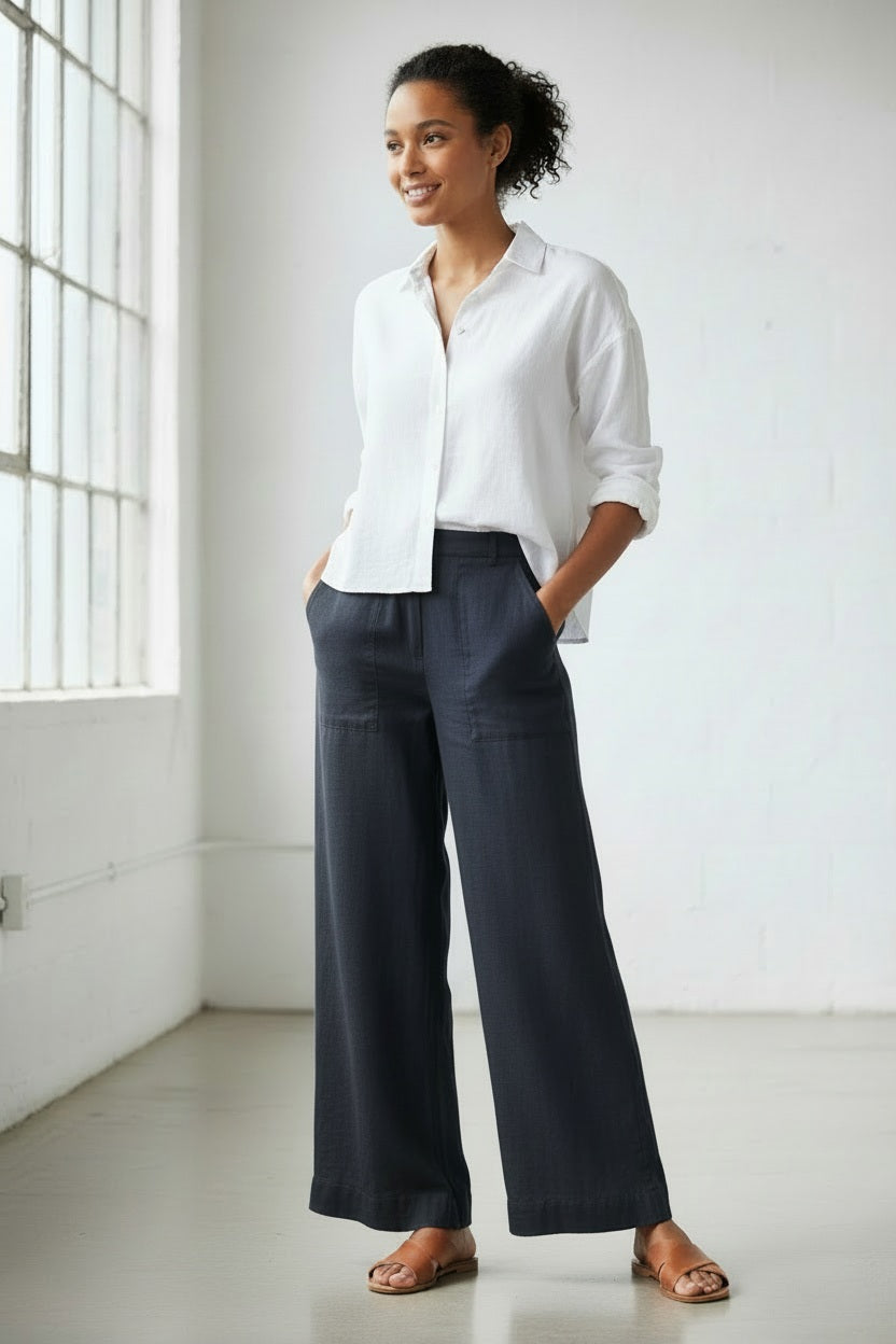 A person stands on a sandy beach wearing a white long-sleeve shirt, FRANK &amp; EILEEN MADELINE Patch Pocket Pull On Wide Leg Linen Pant in Navy, and light-colored sandals. The ocean is visible in the background.
