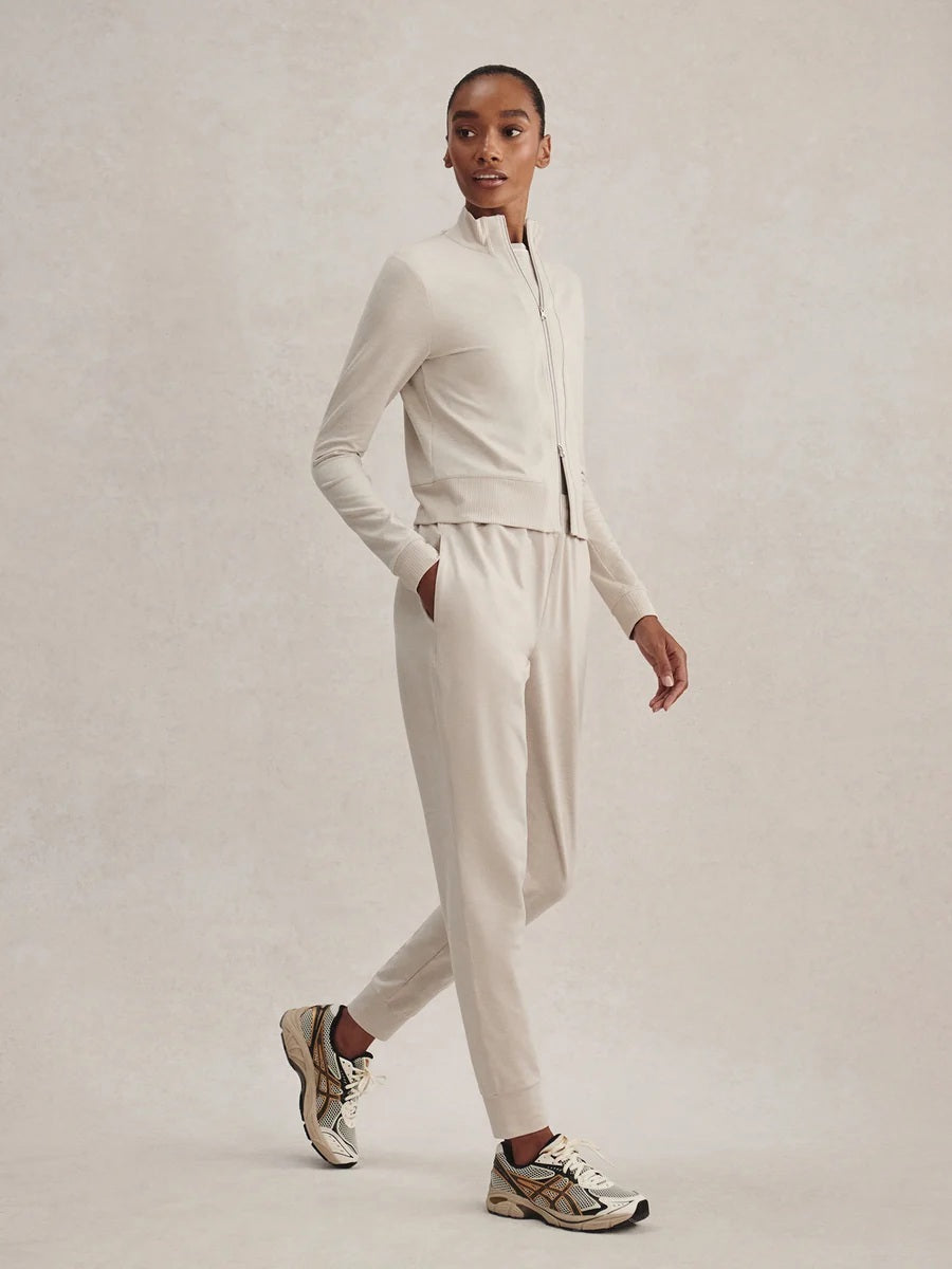 A person wears a light beige Varley Rufus Cropped Zip Through Sweat in Pebble Marl with a high collar and matching pants.